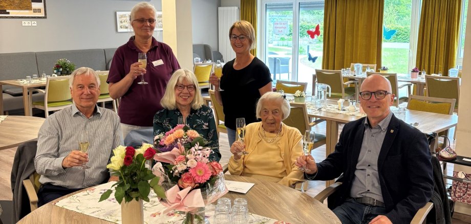 Sechs Personen sitzen an einem Tisch und halten jeweils ein Glas Sekt in der Hand