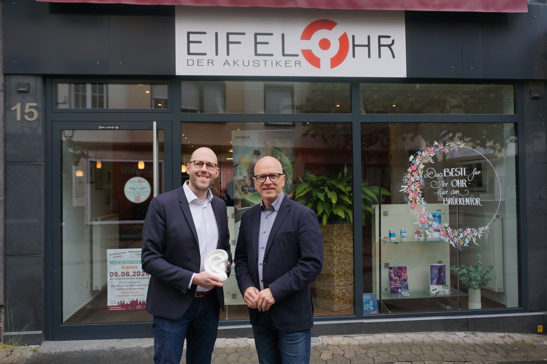 Geschäftsführer Johannes Penzel, Oberbürgermeister Dirk Meid stehen gemeinsam vor dem Schaufenster von Eifelohr