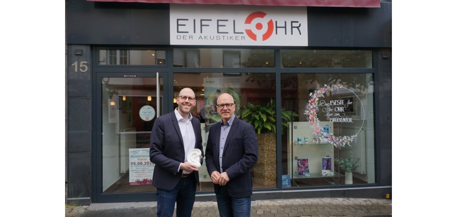 Geschäftsführer Johannes Penzel, Oberbürgermeister Dirk Meid stehen gemeinsam vor dem Schaufenster von Eifelohr