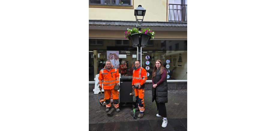 Zwei Mitarbeiter des städtischen Betriebshof und eine Mitarbeiterin der Stadtverwaltung stehen neben einem der neuen Mülleimer