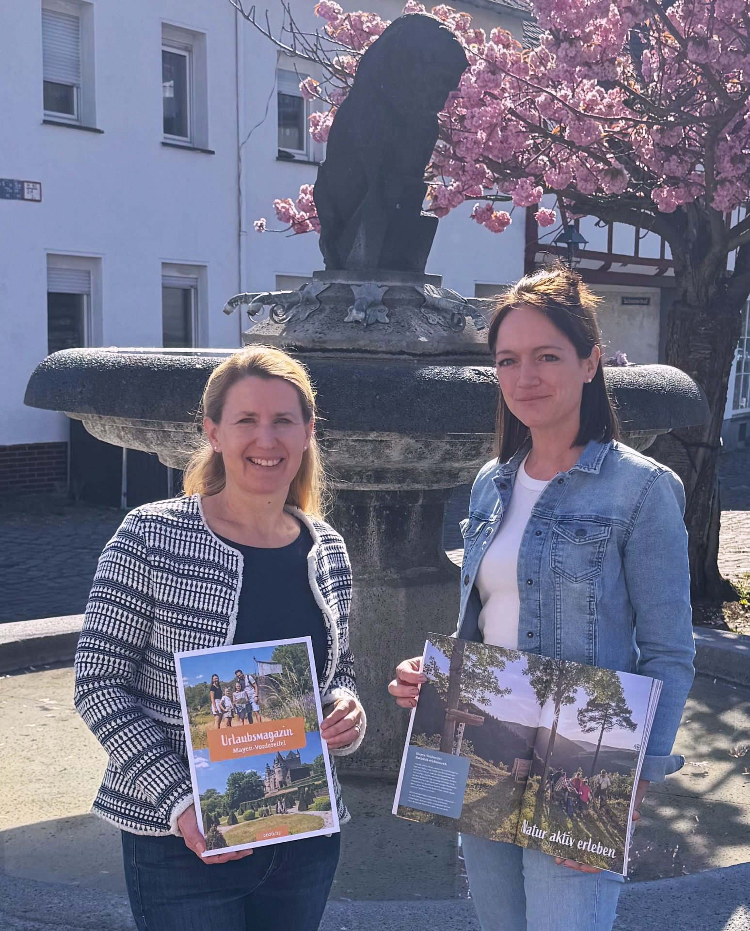 Svenja Schulze-Entrup und Alexandra Burkart stehen draußen vor einem Brunnen und halten das neue Urlaubsmagazin in den Händen 