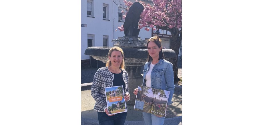 Svenja Schulze-Entrup und Alexandra Burkart stehen draußen vor einem Brunnen und halten das neue Urlaubsmagazin in den Händen 