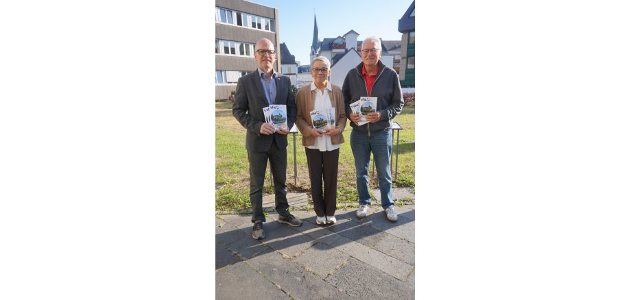 Zwei Männer und eine Frau stehen vor einer Wiese und halten das neue Heft in den Händen