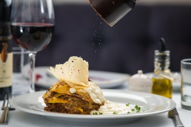 Restaurants ein Teller mit Lasagne, auf die ein Gewürz gemahlen wird. Dazu im Hintergrund ein Glas Rotwein und weitere Gedecke